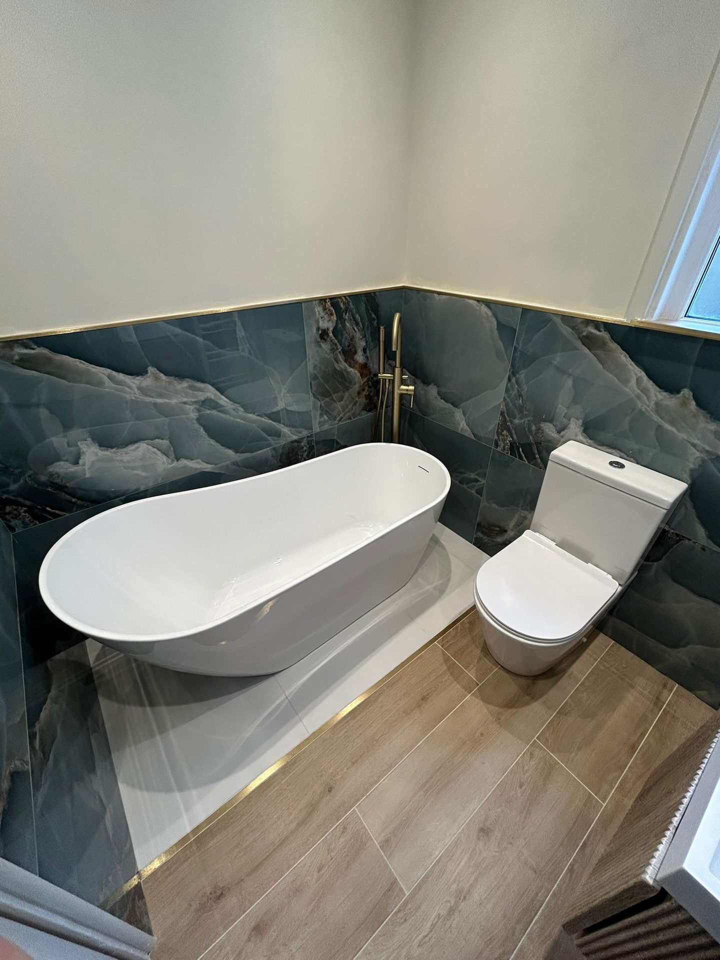 A freestanding white bath on a raised white stone plinth, against walls of blue stone-effect tile finished with a brass trim, on a wood-effect floor.