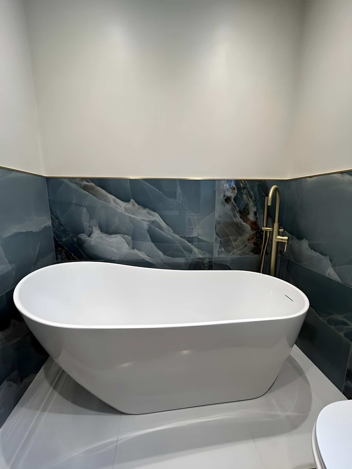 The bath from the doorway: white stone plinth, blue stone-effect tile behind and the brass floor-standing filler in the corner.
