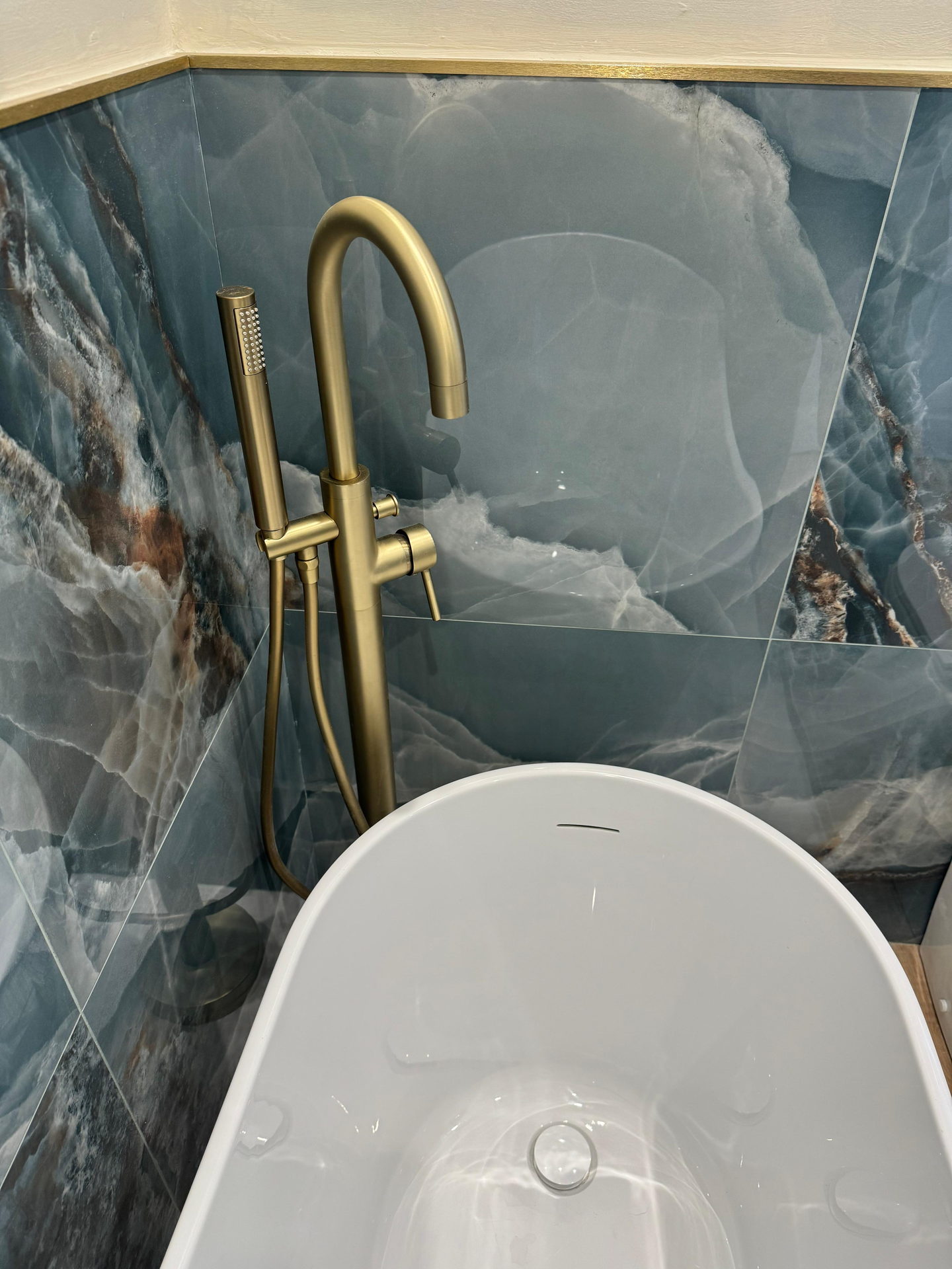 A floor-standing brass bath filler and hand shower against blue stone-effect tile, with the rim of a white bath below.