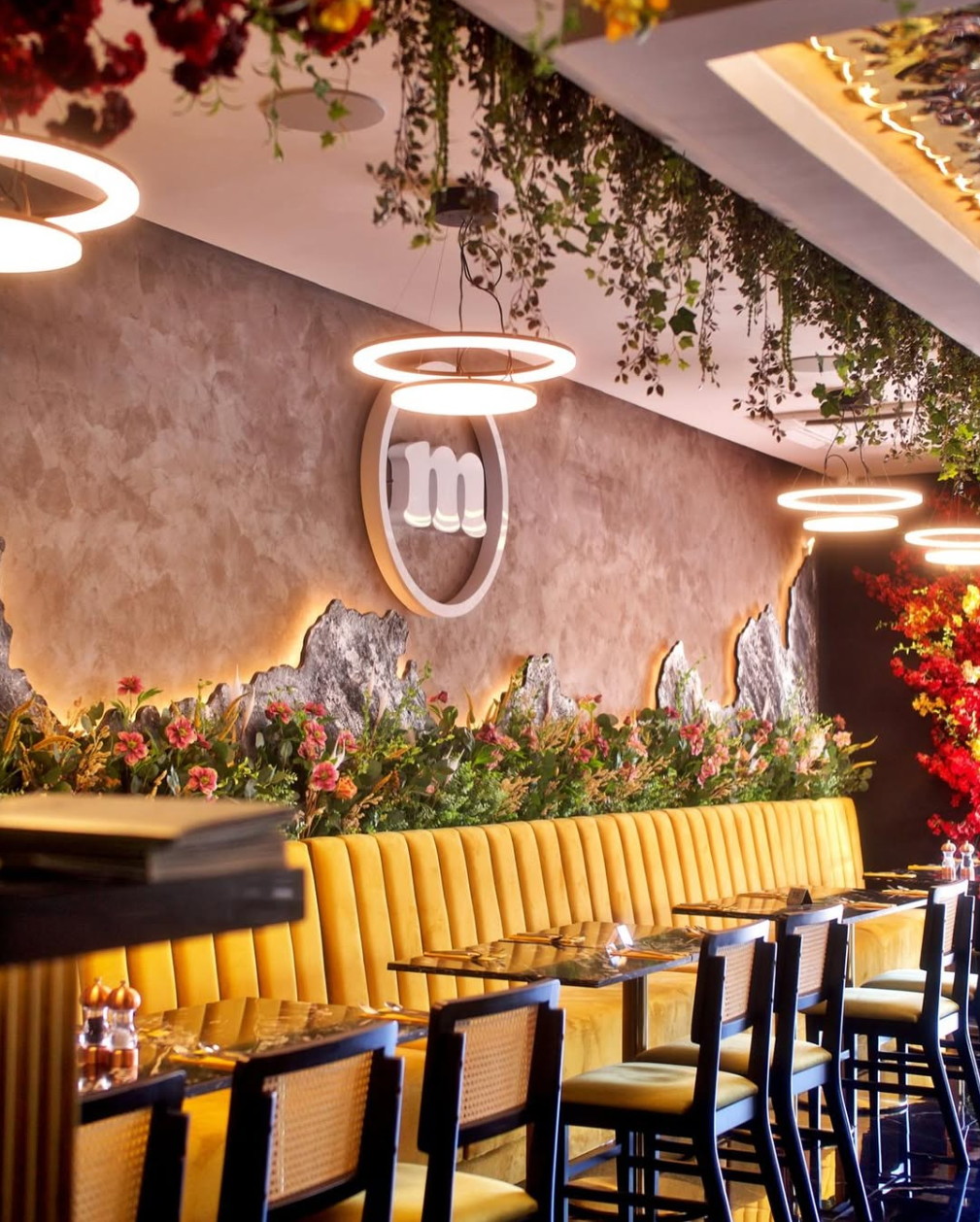 The same restaurant as built: the lit monogram on textured plaster, red flowers overhead among ring lights, and the mustard banquette.