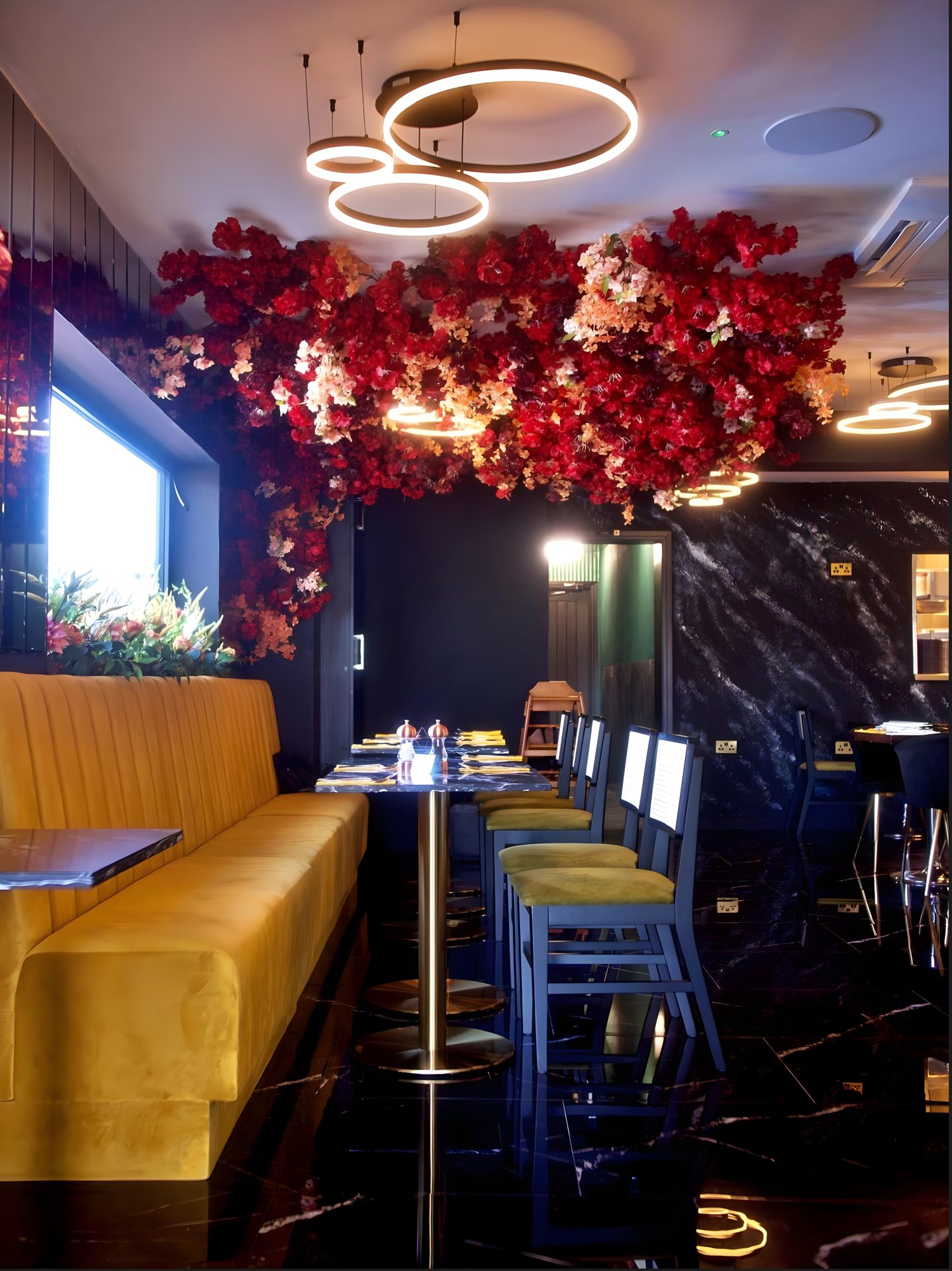 A restaurant with a red floral ceiling installation, gold ring lights, a mustard velvet banquette and a black marble floor.
