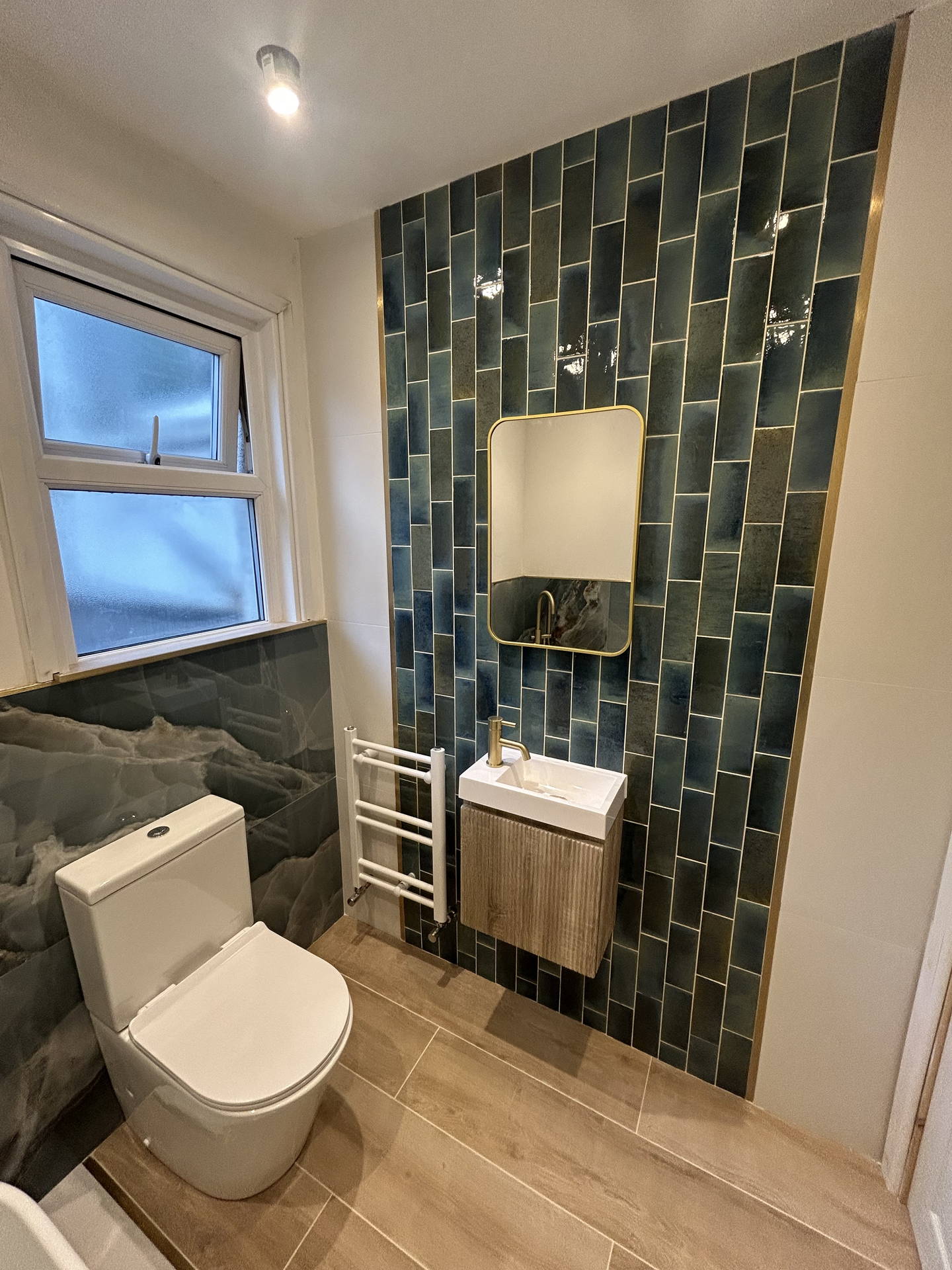 The cloakroom: deep green tile stacked in columns and edged in brass on the wet wall, a brass-framed mirror over a fluted vanity, a white towel rail, and the same blue stone-effect tile to dado height under the window.