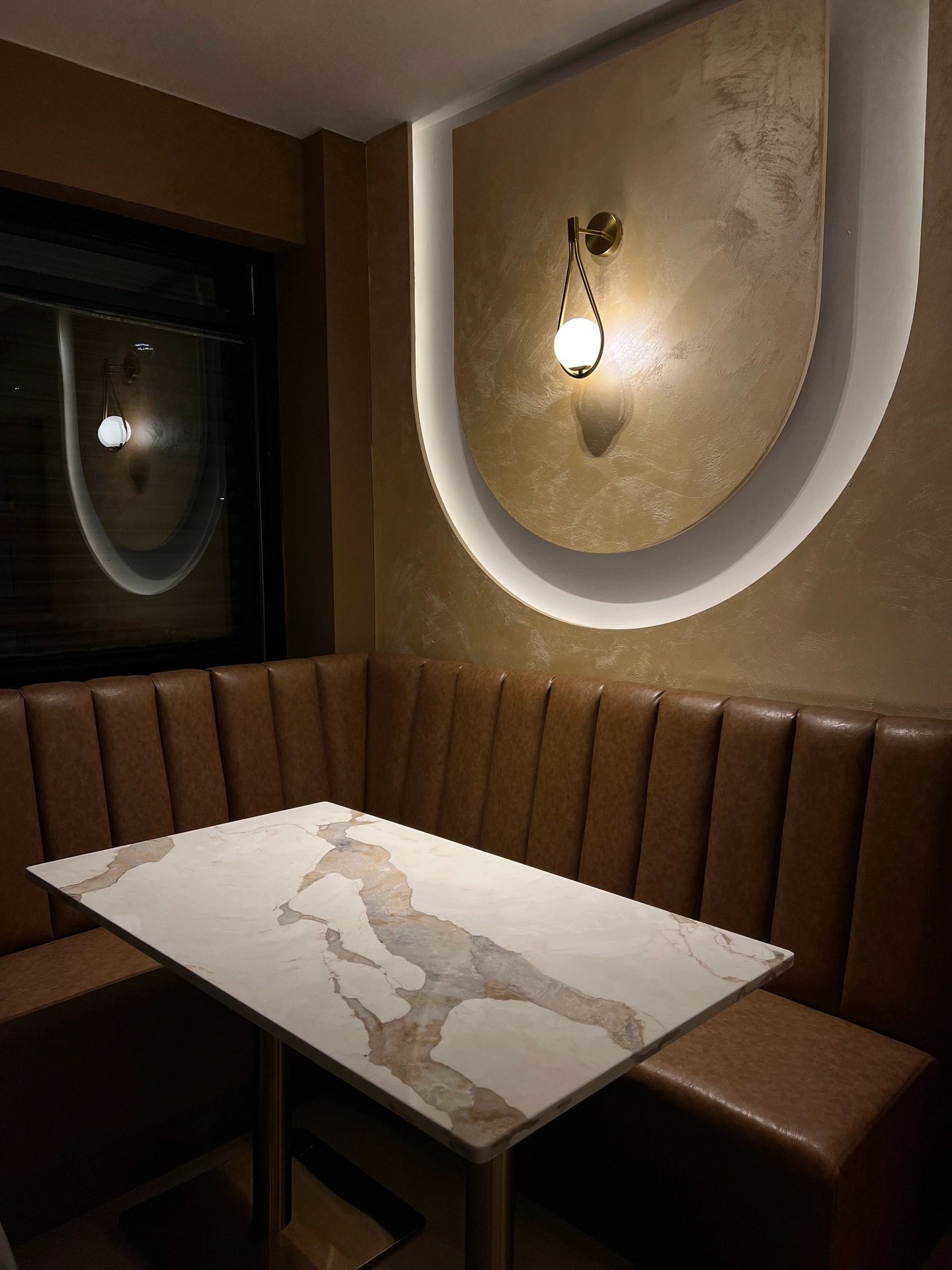 An arched recess in polished plaster lit from within, with a brass wall light, above a fluted leather banquette at Kashmiri Karahi.