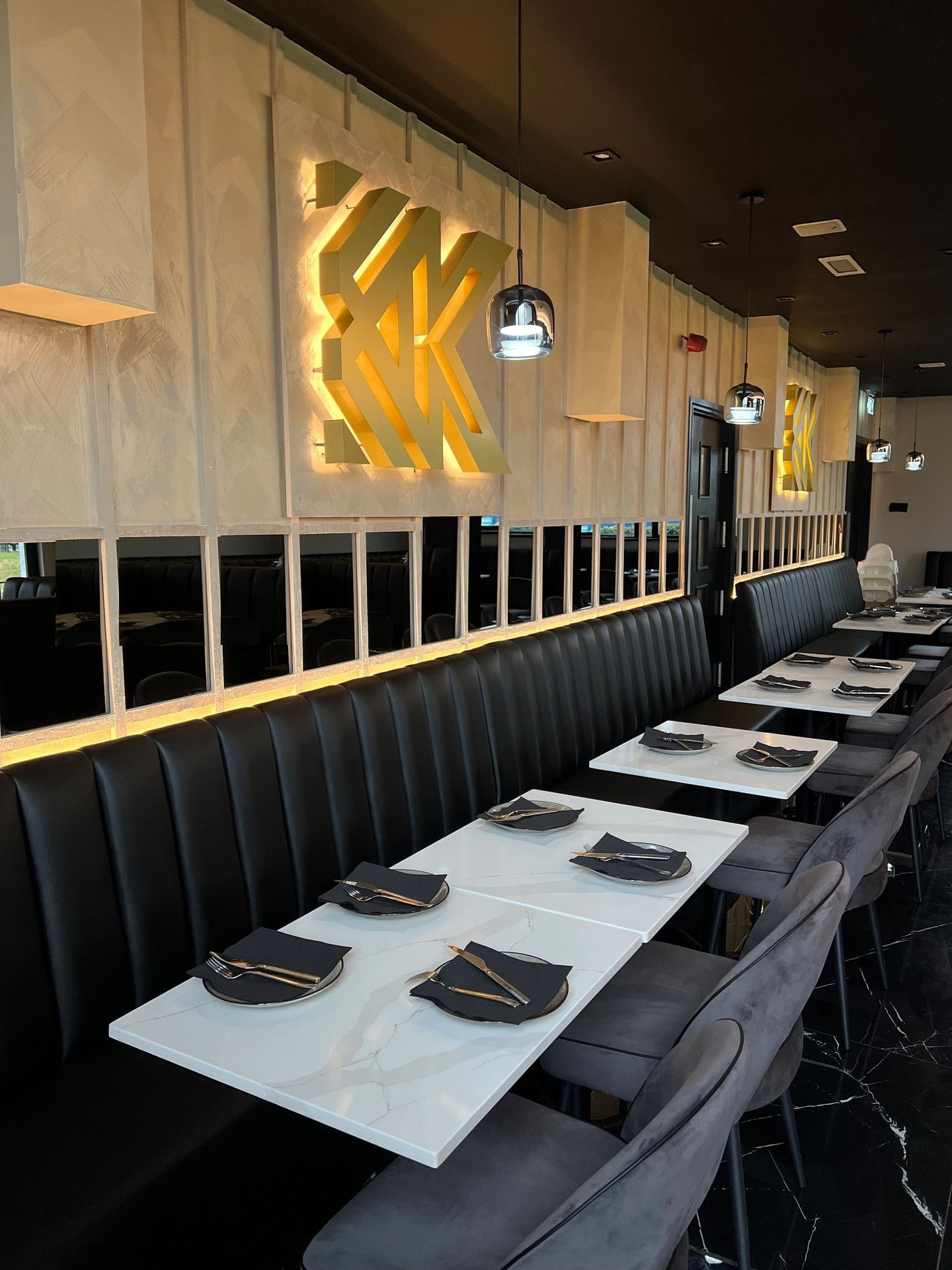 The Kashmiri Karahi dining room in Slough: a backlit gold KK monogram on polished plaster, black fluted leather banquettes, stone-topped tables and a mirrored wall.