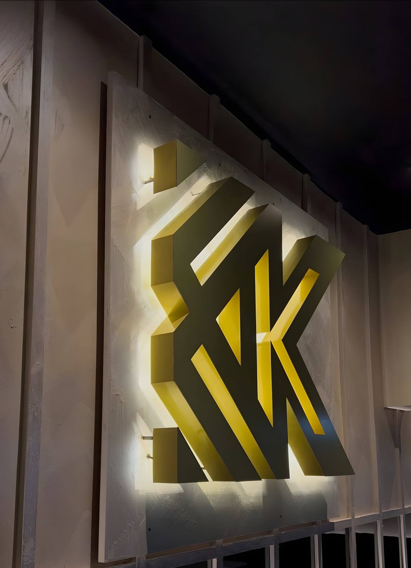 The backlit gold KK monogram, mounted on a polished plaster panel and lit from behind under a black ceiling.