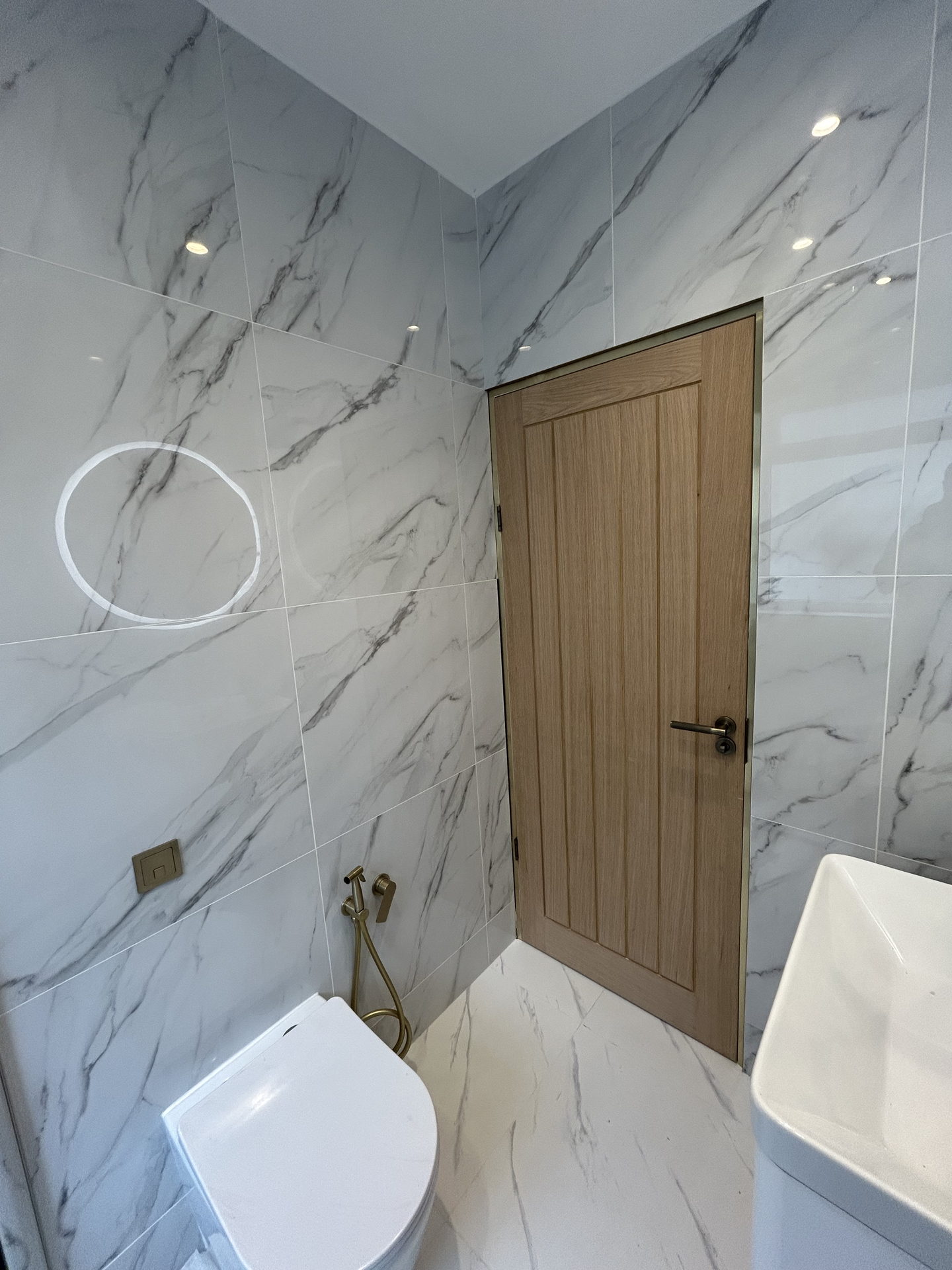 The room from the shower end: white marble-effect tile on every wall, the fluted oak door edged in brass, a wall-hung toilet with a brass hand spray and the edge of the basin.
