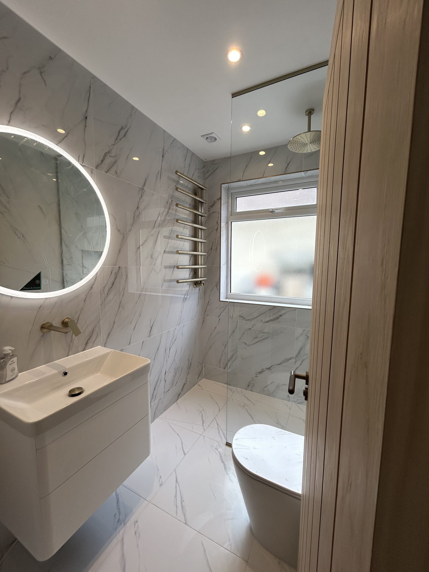 A shower room in white marble-effect tile: a round mirror lit from behind over a wall-hung vanity, a brass towel rail and a walk-in shower behind a glass screen, past a fluted oak door.
