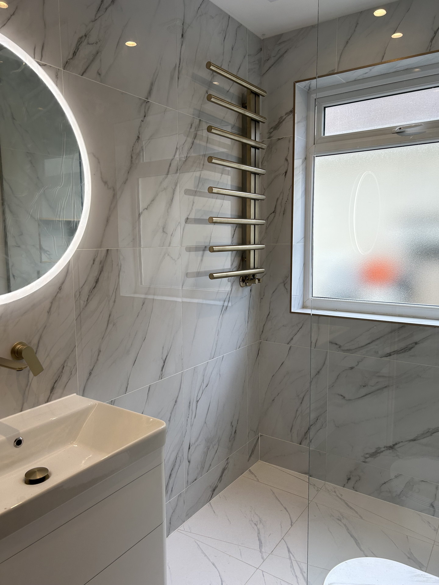 The backlit round mirror over the vanity, the brass towel rail and the frosted window over the shower, on white marble-effect tile.