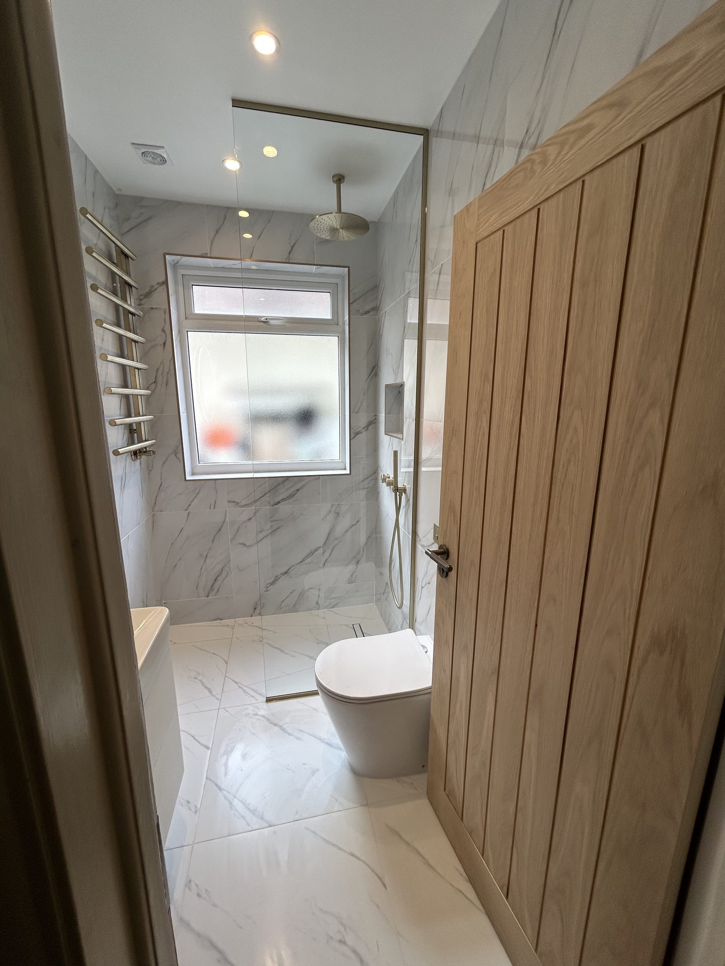 The shower room from the doorway: white marble-effect tile on every wall and the floor, a brass towel rail and a fixed glass screen, past a fluted oak door.
