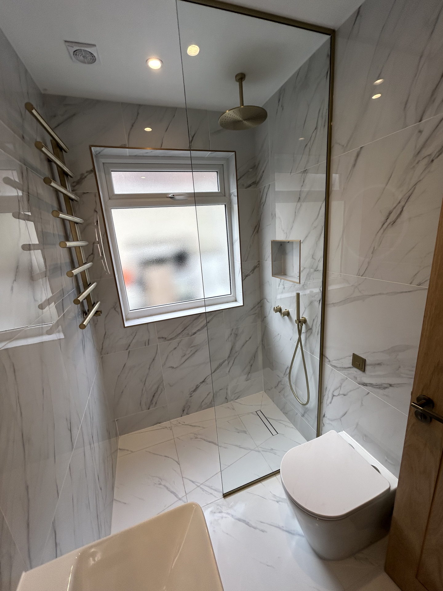 The walk-in shower behind a fixed glass screen: a brass rain head dropped from the ceiling, a frosted window, a recessed niche and a brass hand shower.