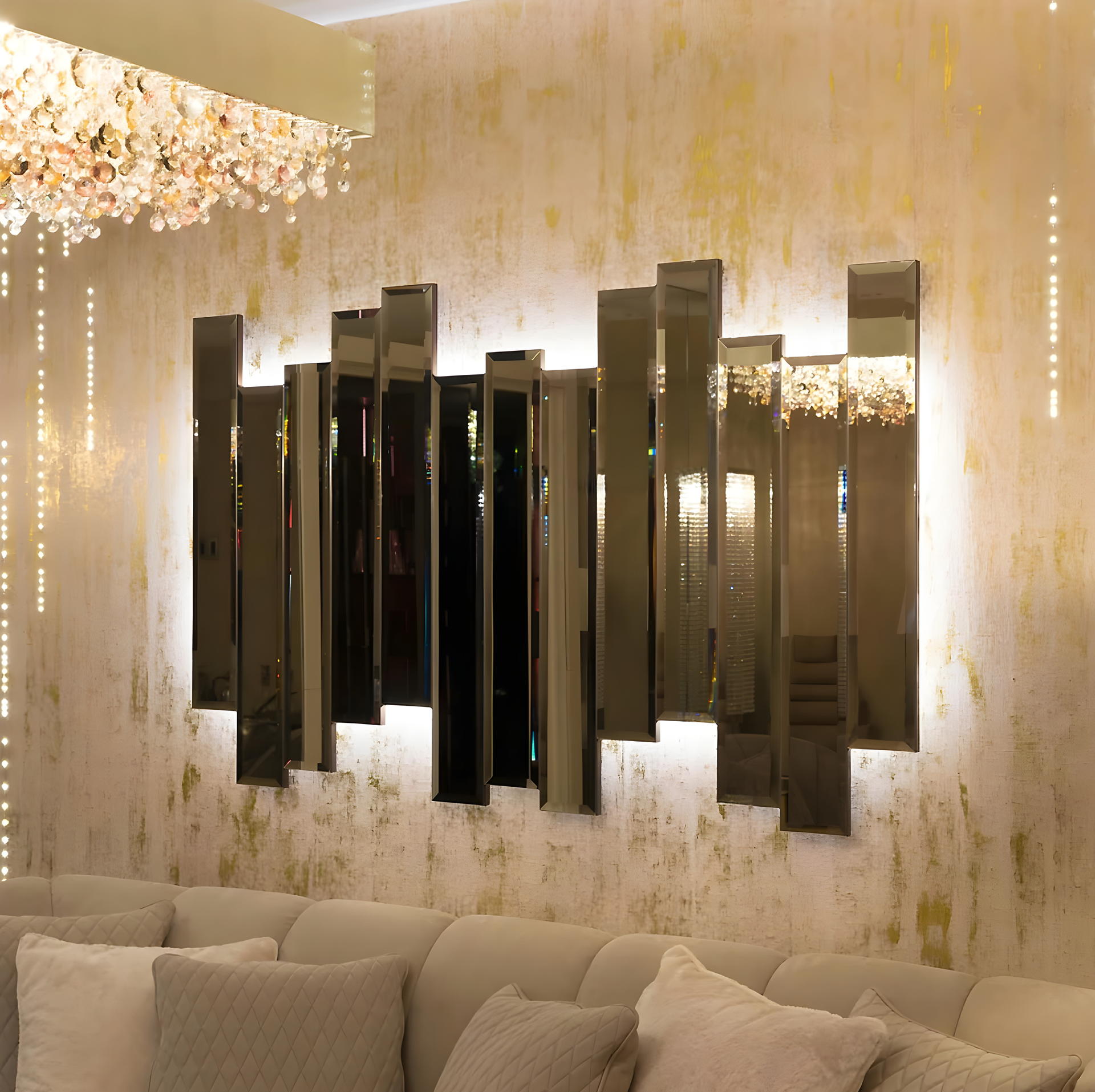 Bevelled bronze mirror panels set at staggered heights and lit from behind, on a wall of gold leaf over plaster, under a chandelier of crystal drops, above a cream sofa.