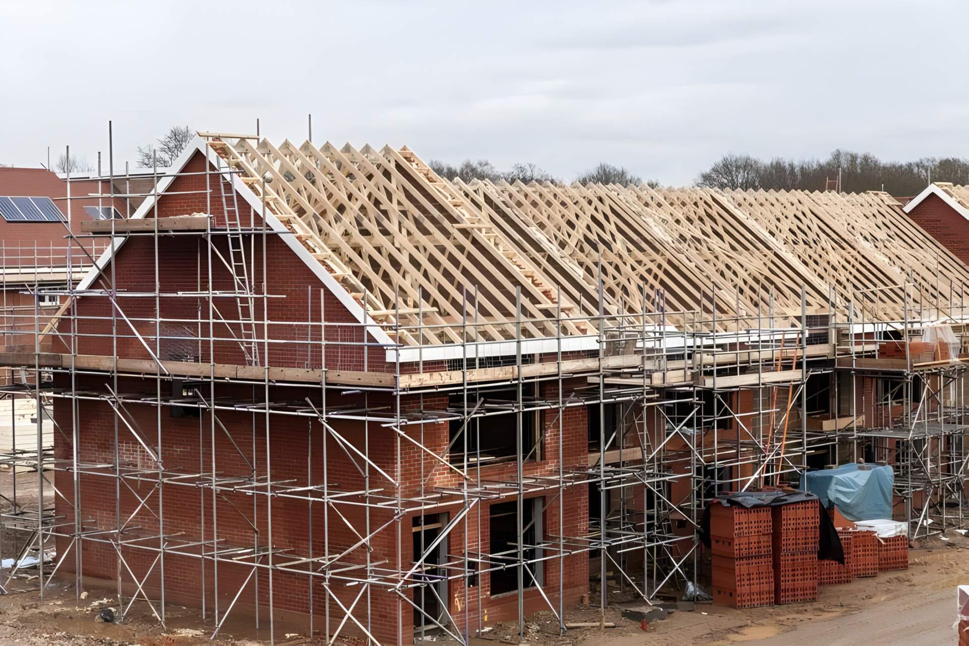 A new build in progress: a brick shell under scaffold with timber roof trusses and packs of bricks.