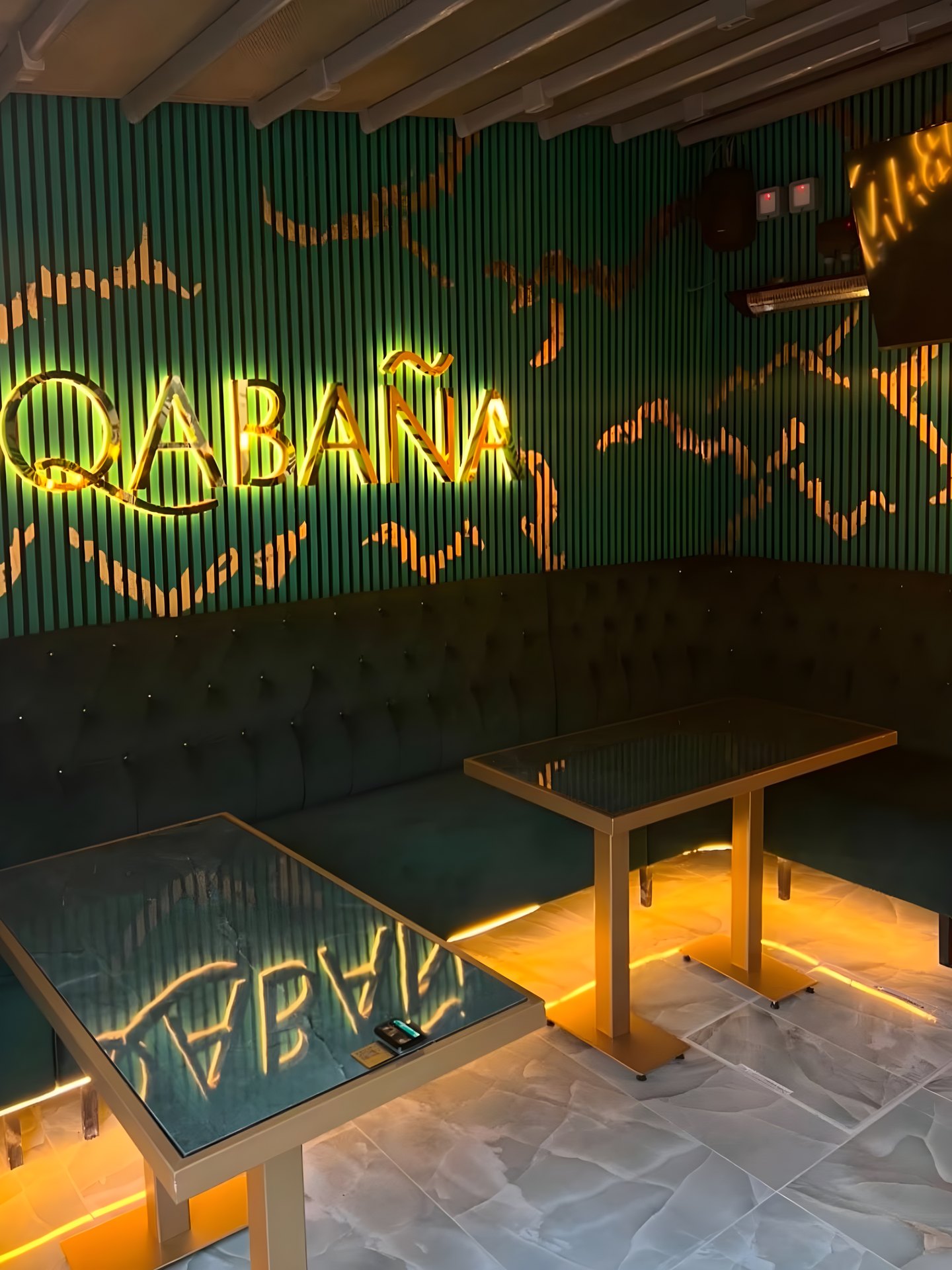 The Qabaña wordmark on deep green fluted panelling above a buttoned green banquette, with brass-framed dark-glass tables lit from beneath.