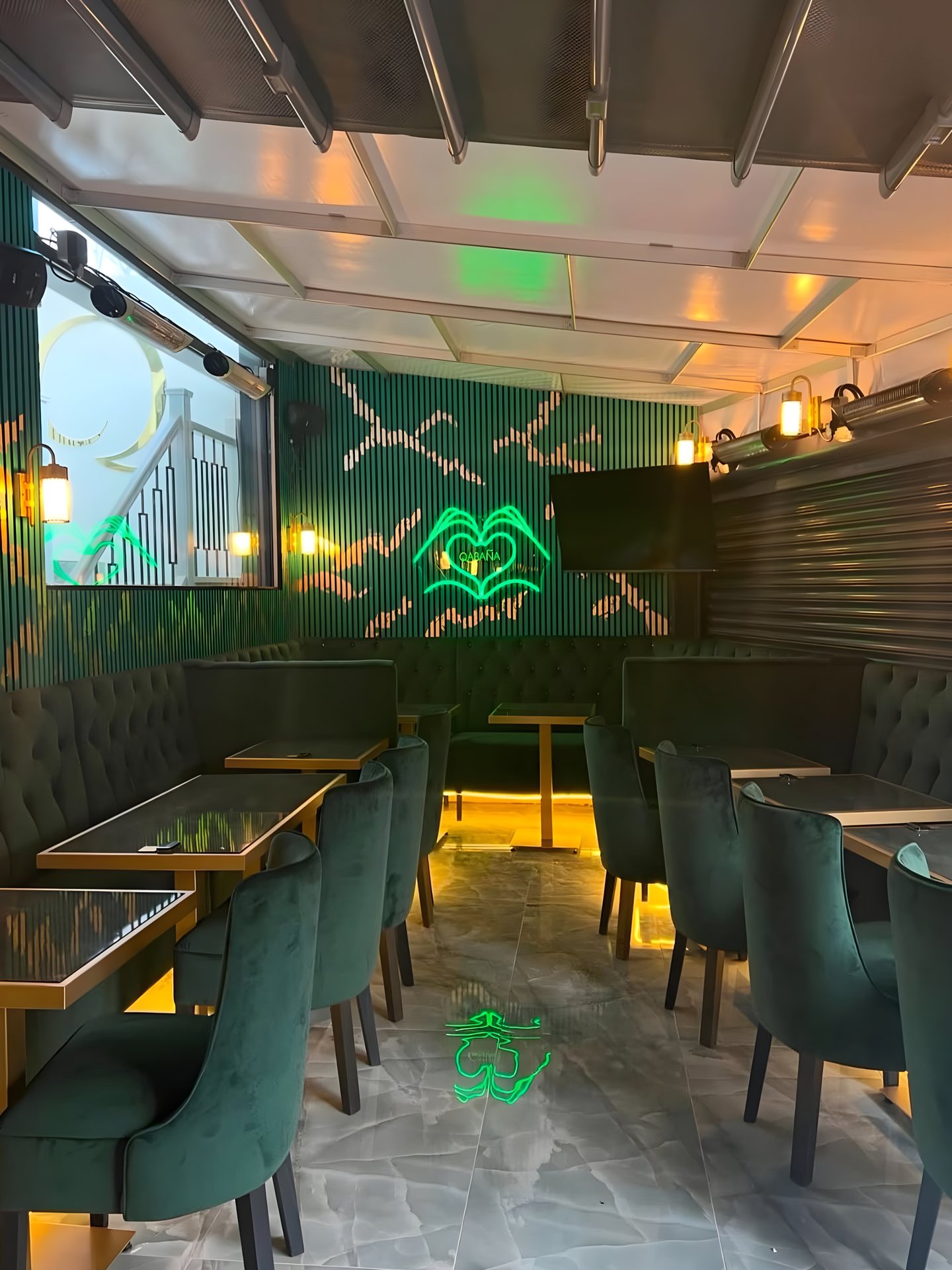 The seating area at Qabaña from the corner: green chairs at brass-toned tables, the buttoned banquette along the panelled walls, a green neon artwork and wall lanterns.