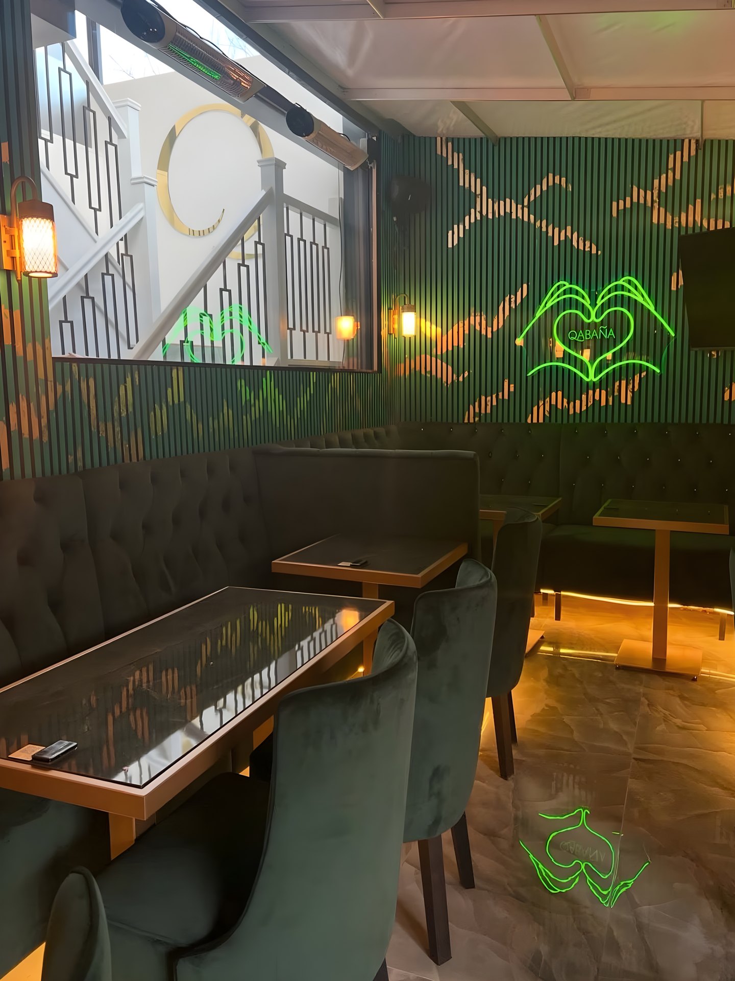 Looking towards the stair and window with a gold ring light, past the buttoned banquette, green chairs, a lantern on the panelling and the green neon artwork.