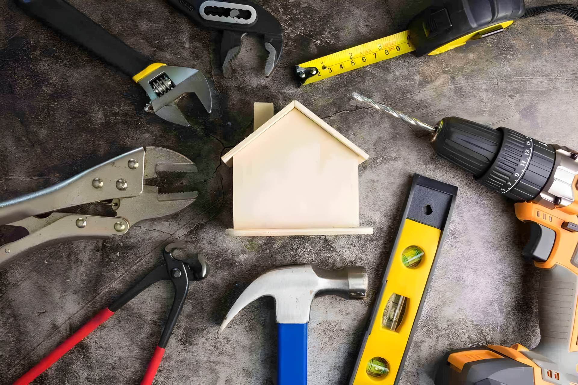 Hand tools laid out around a small wooden house: a spirit level, a tape measure, a hammer, a drill and wrenches.